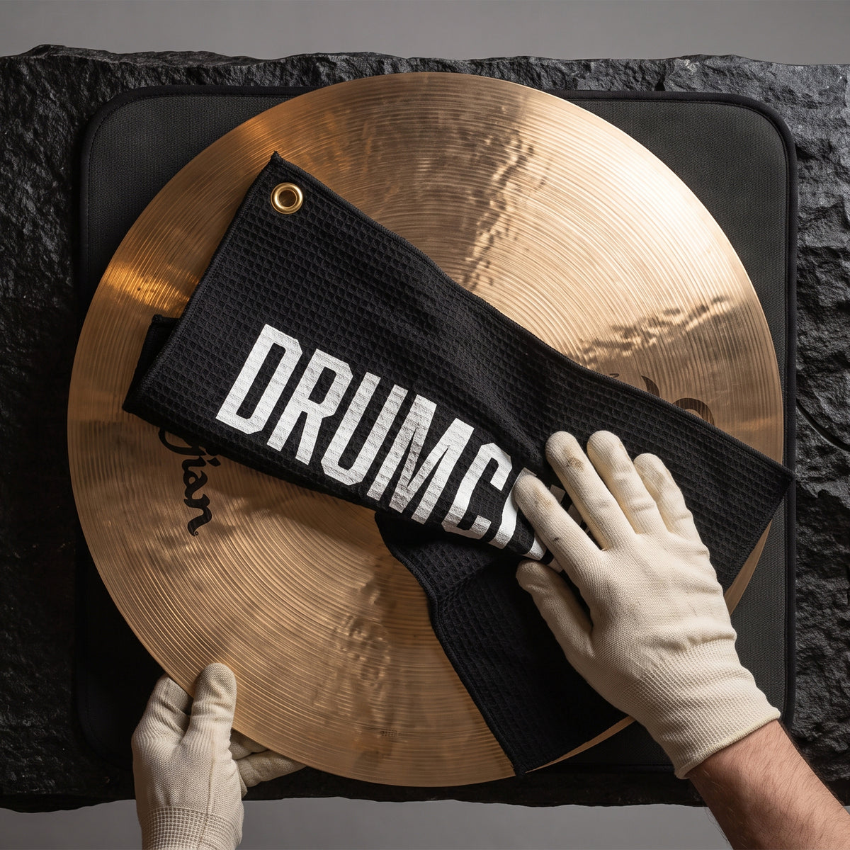 A black microfiber cleaning cloth with white 'DRUMCRAFT' lettering is being wiped onto a gold cymbal by gloved hands, set against a dark stone background.